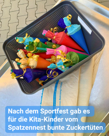 Ein grauer Korb, der überquillt von farbenfrohen „Schultüten“ oder "Zuckertüten". Die Tüten präsentieren sich in verschiedenen leuchtenden Farben wie Blau, Rot, Pink, Grün und Orange, jede gekrönt von Tissue- oder Crêpe-Papier und mit Bändern verziert, was ihrem Festcharakter zusätzlichen Glanz verleiht.  Der Korb steht auf einer metallischen Oberfläche, darunter ist eine teilweise sichtbare, gefaltete Textilie oder Tragetasche mit blauen Streifen und Text zu erkennen. Auf dem Bild steht: „Nach dem Sportfest gab es für die Kita-Kinder vom Spatzennest bunte Zuckertüten.“